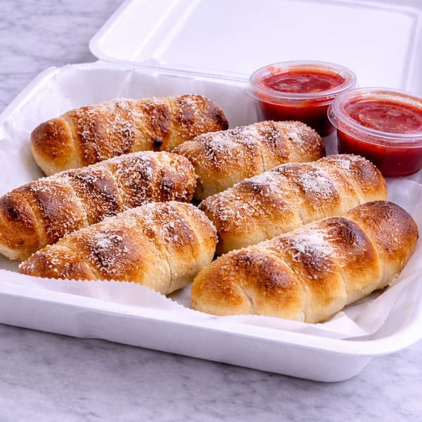Six golden-brown garlic knots, sprinkled with Parmesan cheese, in a white takeout container with two cups of marinara sauce.